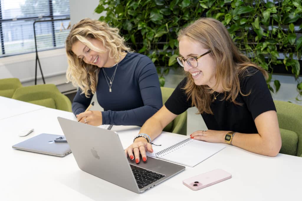 Twee vrouwen zitten samen lachend aan een vergadertafel, werkend met een laptop en notitieboek, met groene plantenwand op de achtergrond.